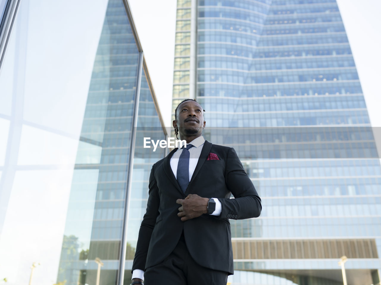 Serious african american male entrepreneur wearing elegant suit walking along glass office building in city center and looking away