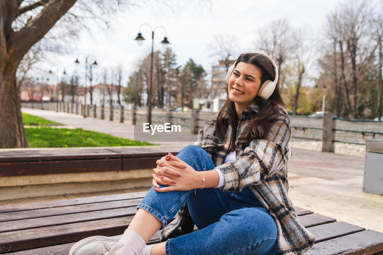 sitting, adult, women, one person, young adult, smiling, emotion, happiness, bench, lifestyles, casual clothing, relaxation, leisure activity, nature, architecture, city, park bench, spring, clothing, tree, photo shoot, person, park, jeans, female, long hair, city life, autumn, positive emotion, park - man made space, hairstyle, portrait photography, cheerful, portable information device, winter, portrait, enjoyment, outdoors, technology, looking, smartphone, wireless technology, sky, brown hair, three quarter length, communication, day, listening, teeth, teenager, mobile phone, smile, footwear, denim, fashion, telephone, contemplation, seat