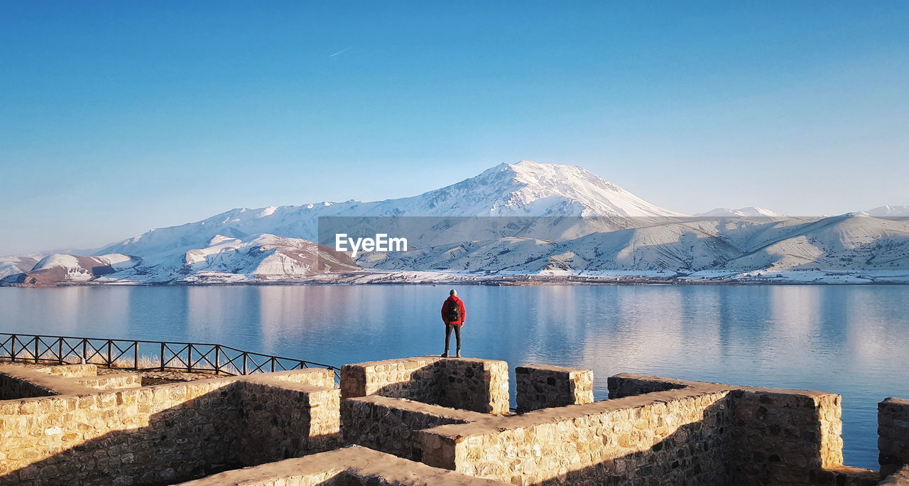 Rear view of man standing on stone wall by lake during winter