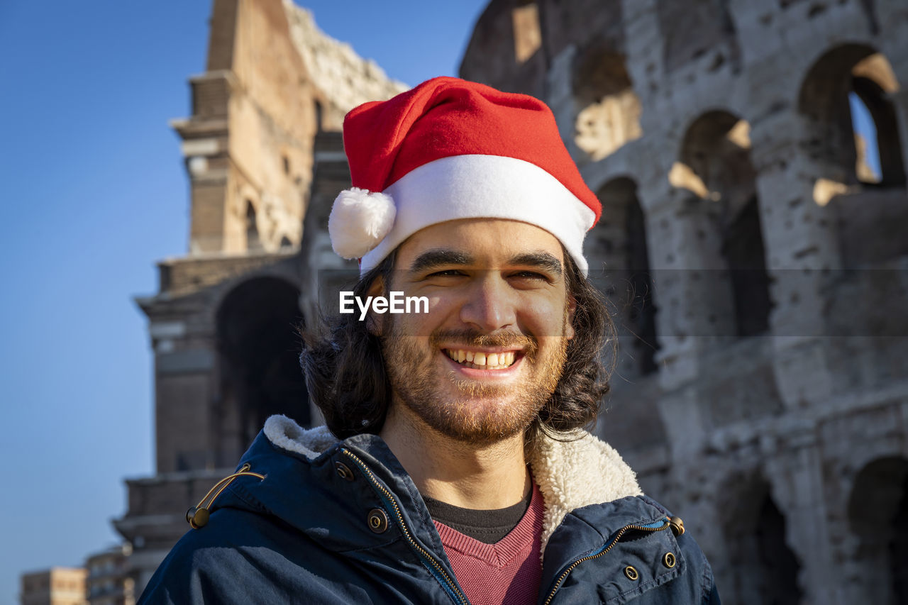 Smiling young man with a santa hat in front of the colosseum. christmas roman holiday concept.