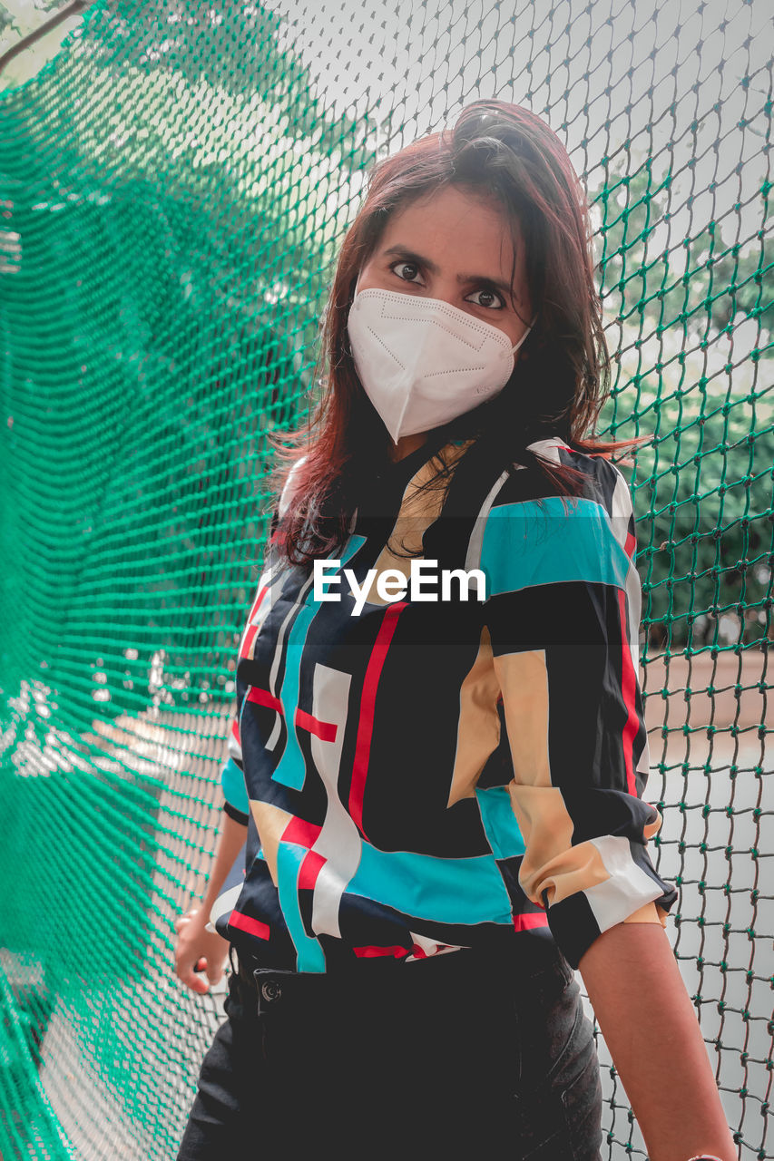 Portrait of woman wearing flu mask standing against net outdoors
