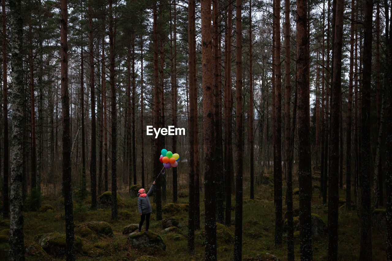 Woman holding balloons standing amidst trees in forest