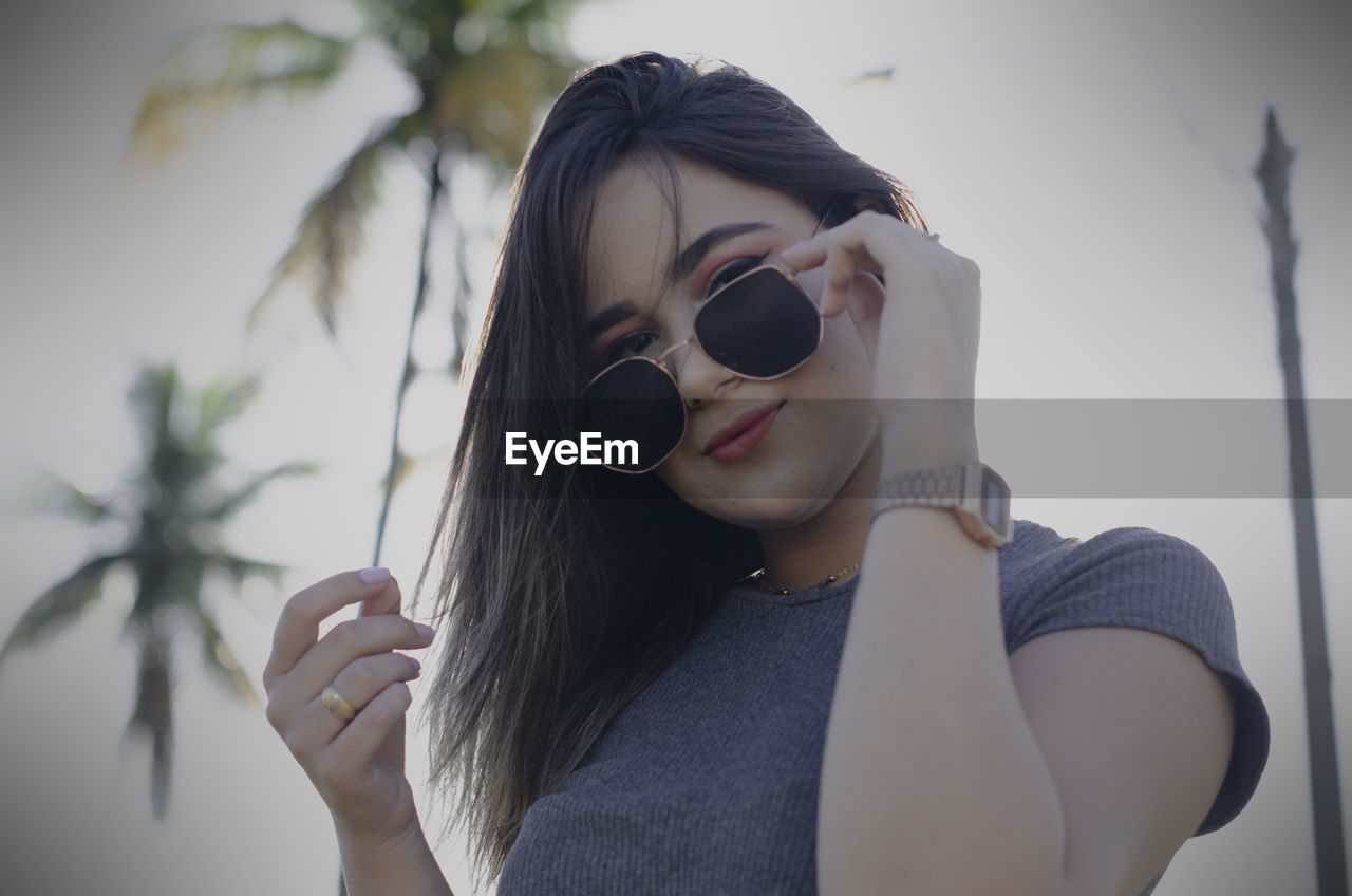 PORTRAIT OF YOUNG WOMAN WEARING SUNGLASSES HOLDING OUTDOORS