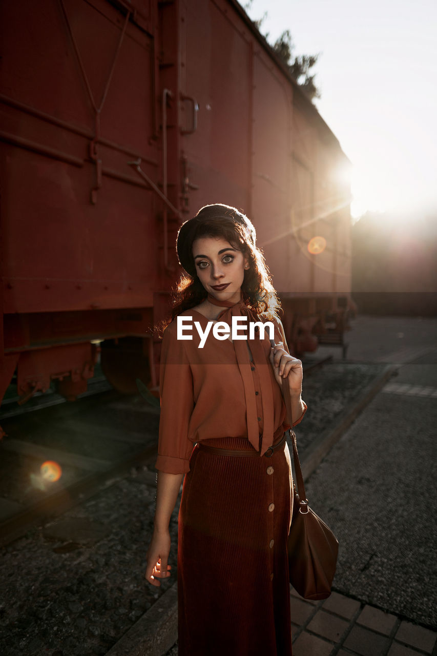 Dark haired female totally wearing terracotta clothes in vintage style standing near terracotta car train and holding open book in hands having dreaming look