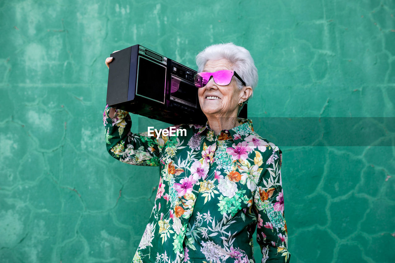 Cool elderly female in colorful trendy blouse and sunglasses carrying record player and enjoying music against green wall