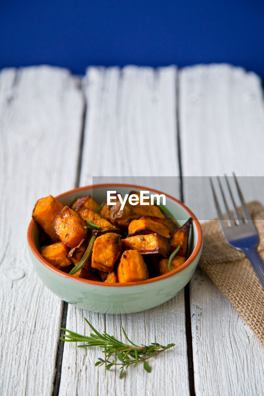 Close-up of bowl of oven roasted sweet potato cubes with fresh herbs on rustic white wooden table