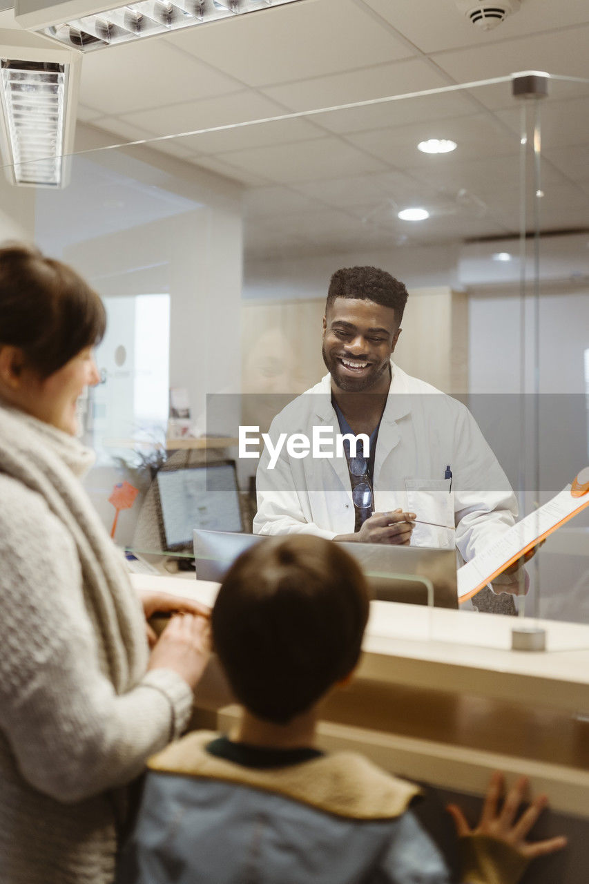 Smiling male receptionist talking to mother and son through transparent shield in clinic