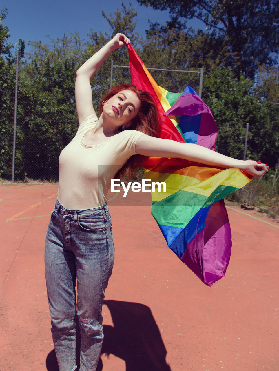 Young woman holding rainbow flag on basketball court against trees