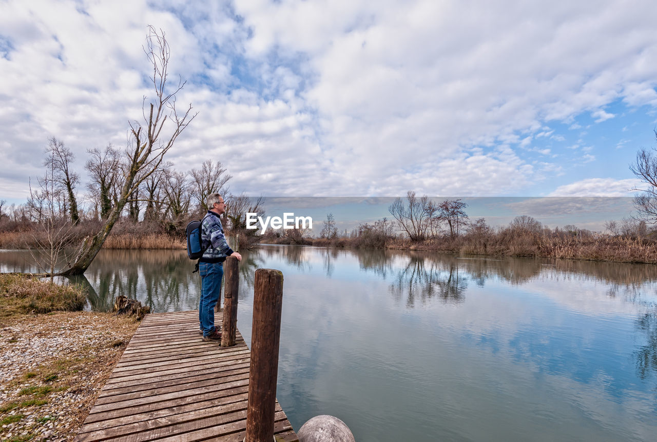 Side view of mature man with backpack standing on pier over lake against cloudy sky