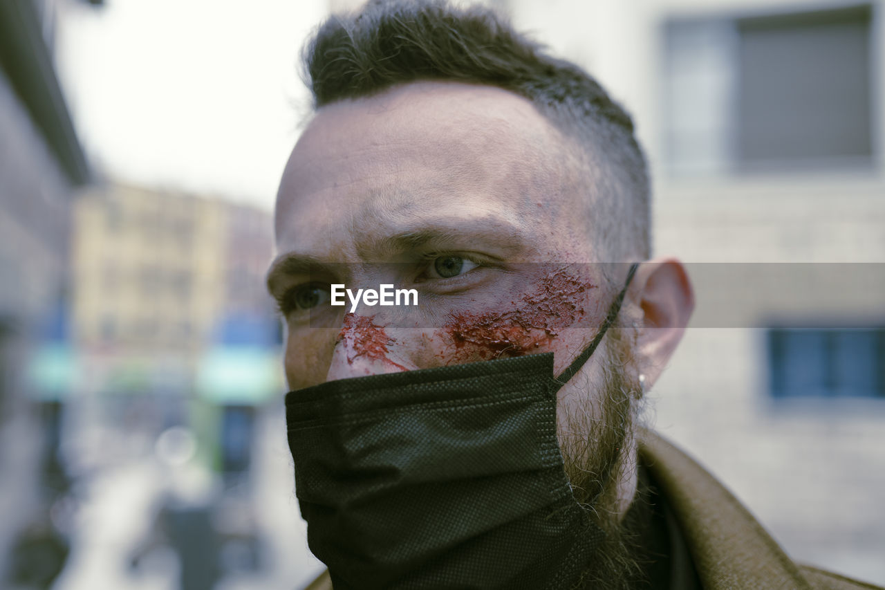 Side view hipster man with beard, cinema makeup and mask posing on the street and looking at camera