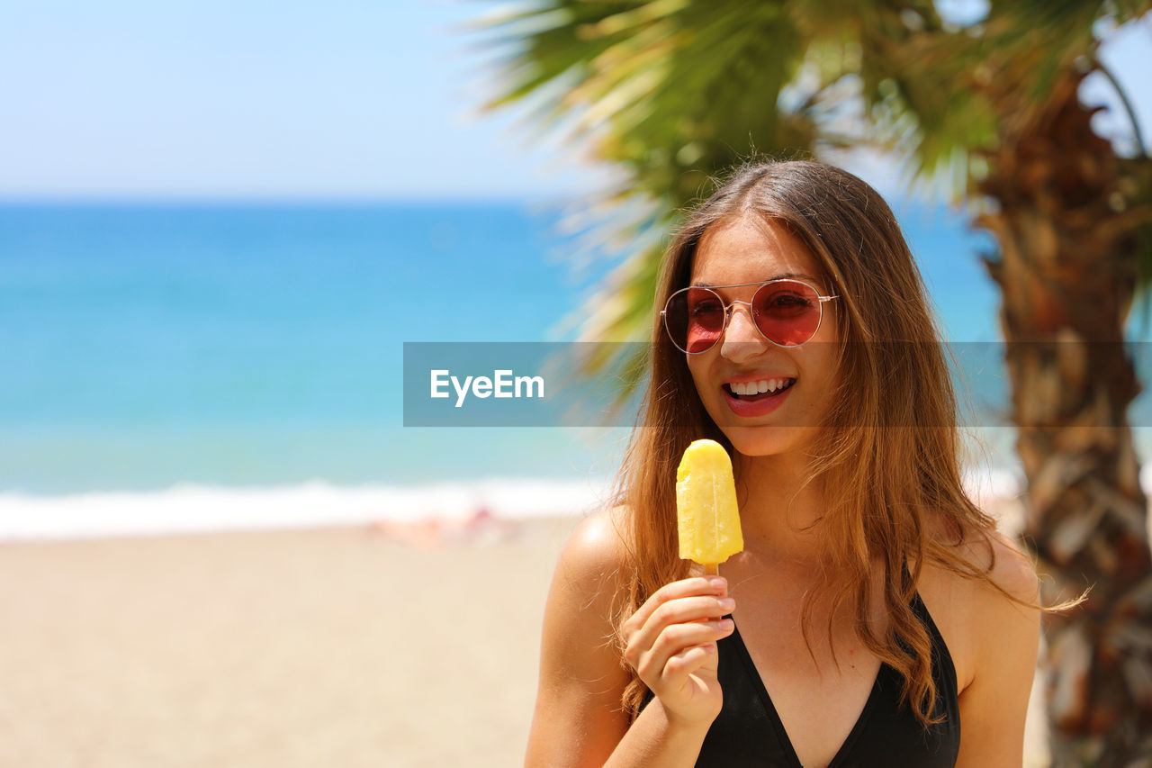 Young woman in sunglasses eating popsicle while standing at beach during sunny day