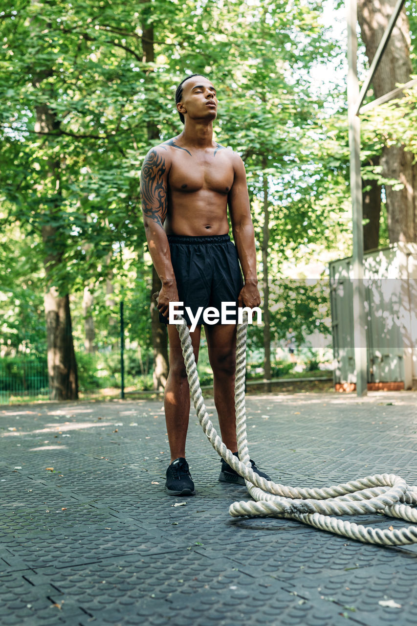 Full length of shirtless man holding rope while standing against trees