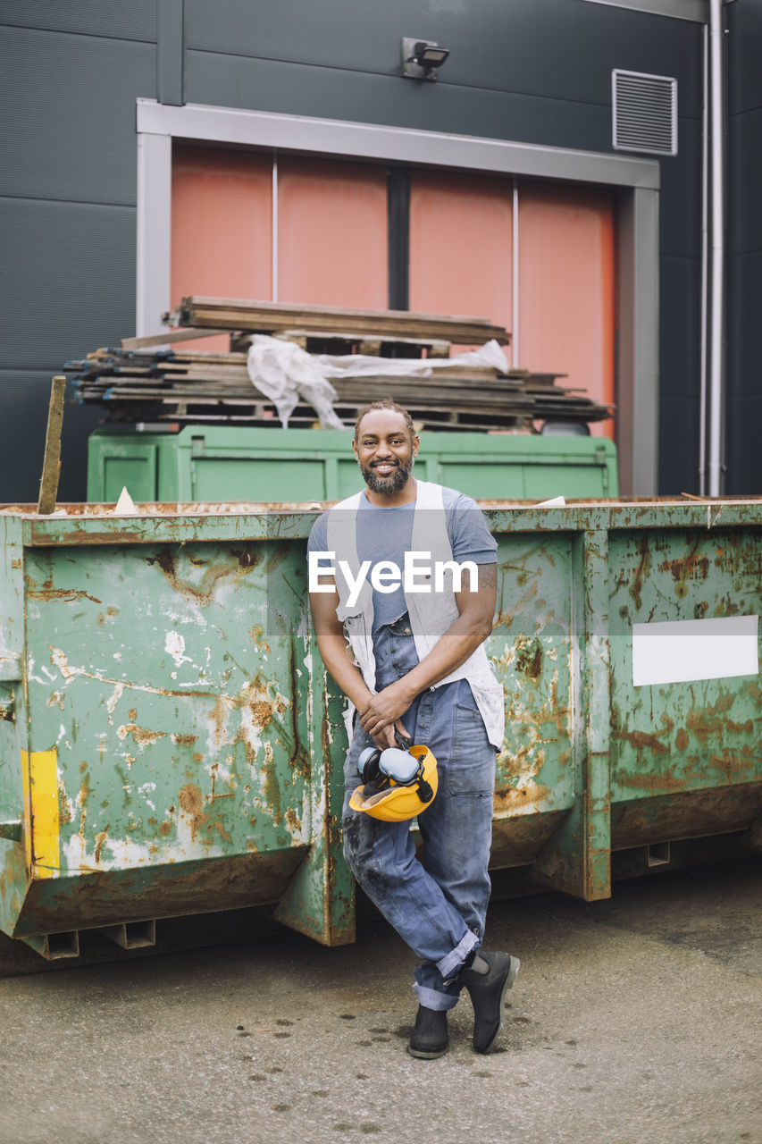 Full length of smiling male construction worker with hardhat leaning on metal container at site