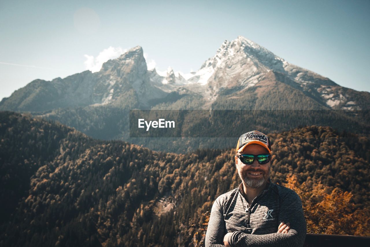 PORTRAIT OF SMILING MAN STANDING BY MOUNTAIN AGAINST SKY