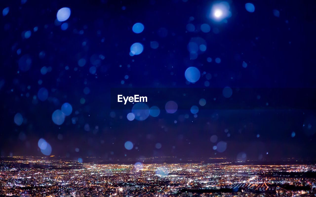 Snowfall over illuminated cityscape against sky at night