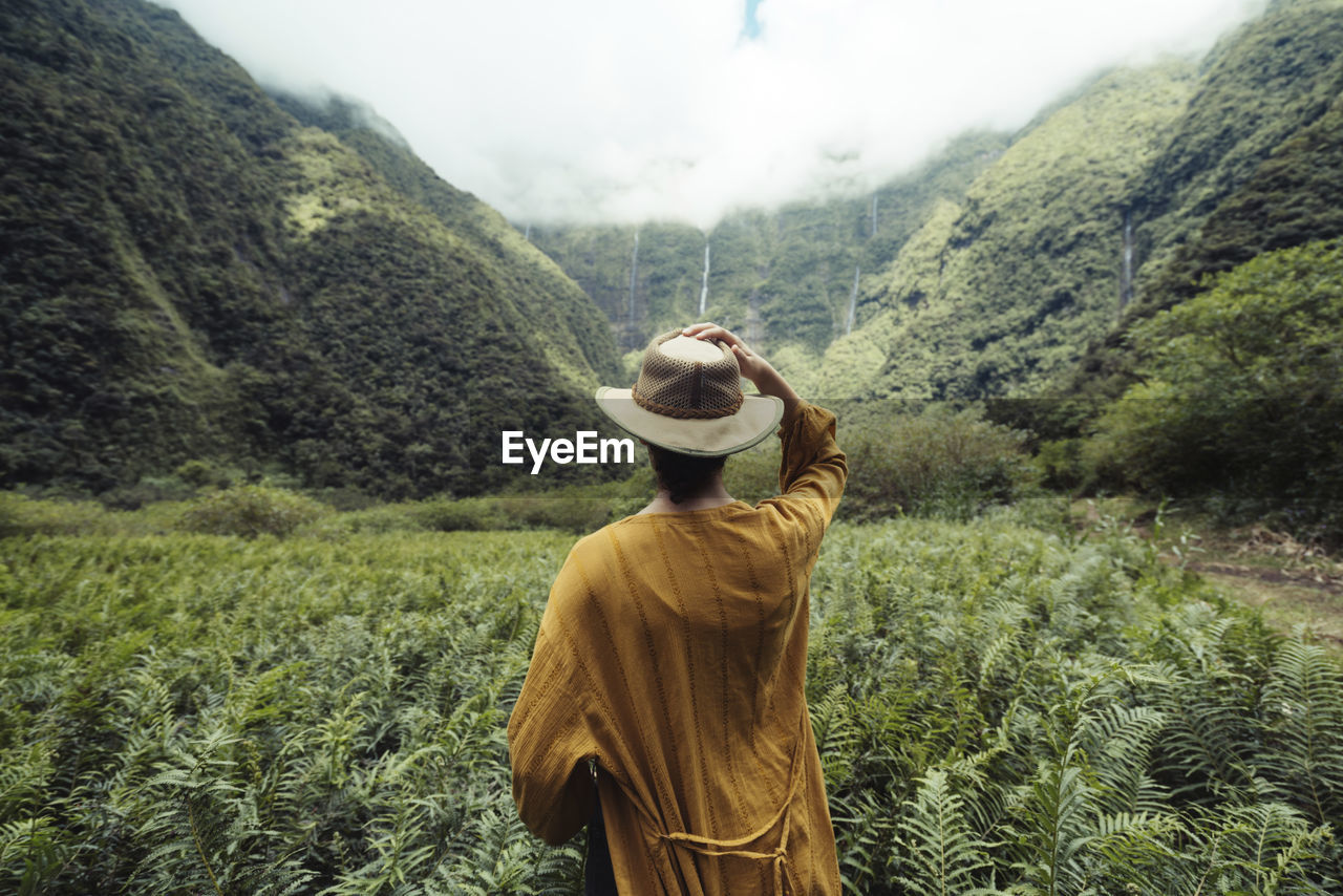 Rear view of woman wearing hat standing against cloudy sky in forest