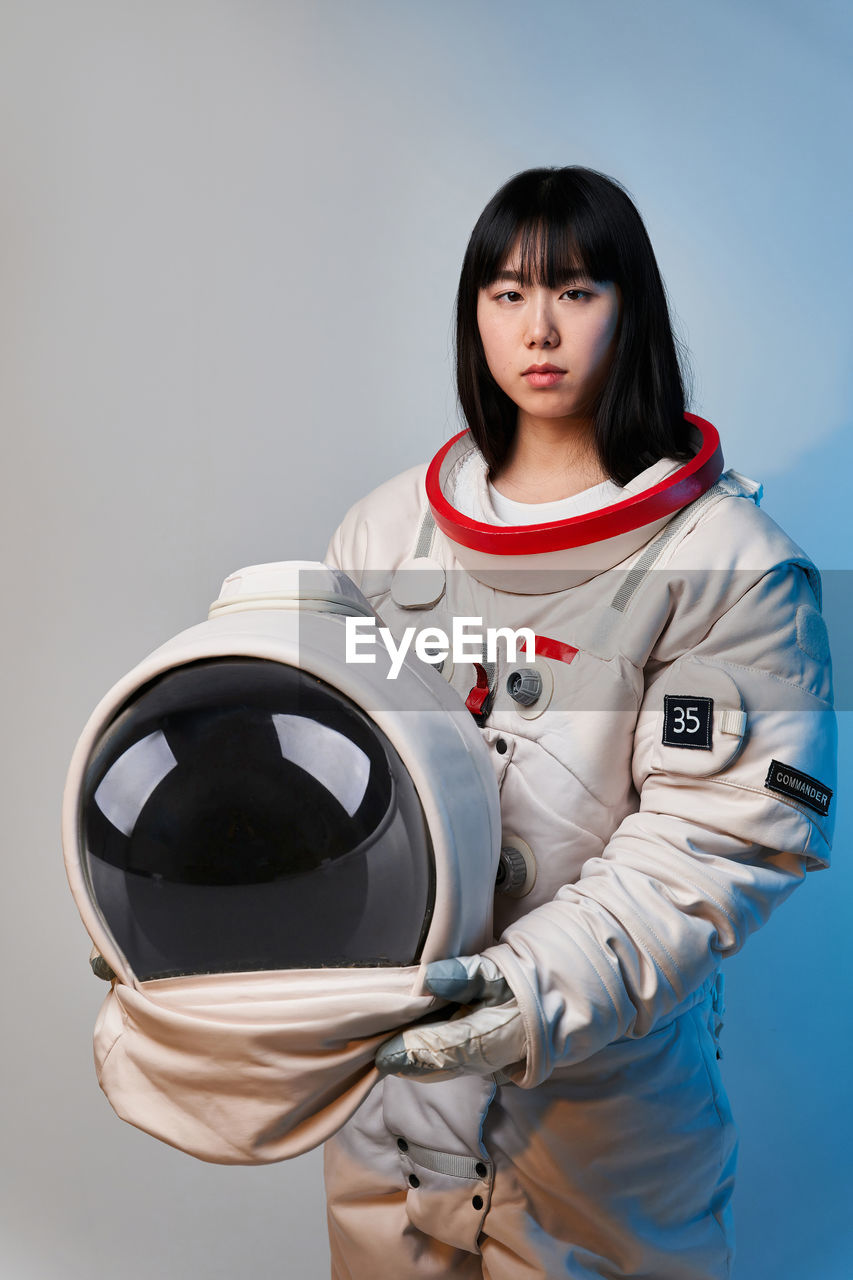 Young asian woman in spacesuit holding helmet and looking at camera under blue light before mission against gray background