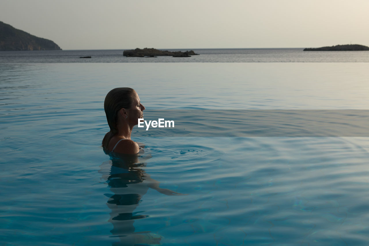 Woman in infinity pool by sea during sunset