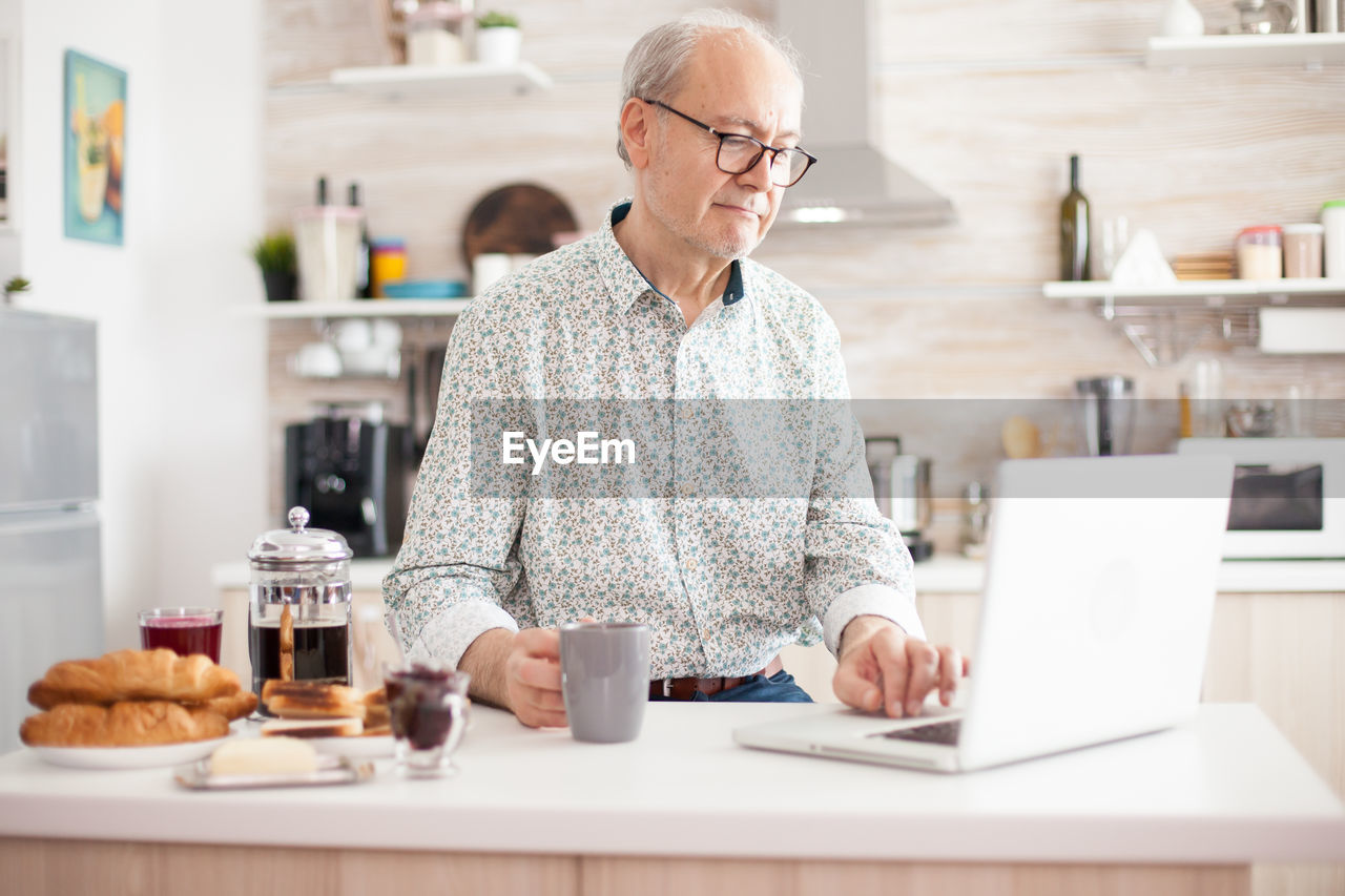Senior man holding coffee cup while using laptop