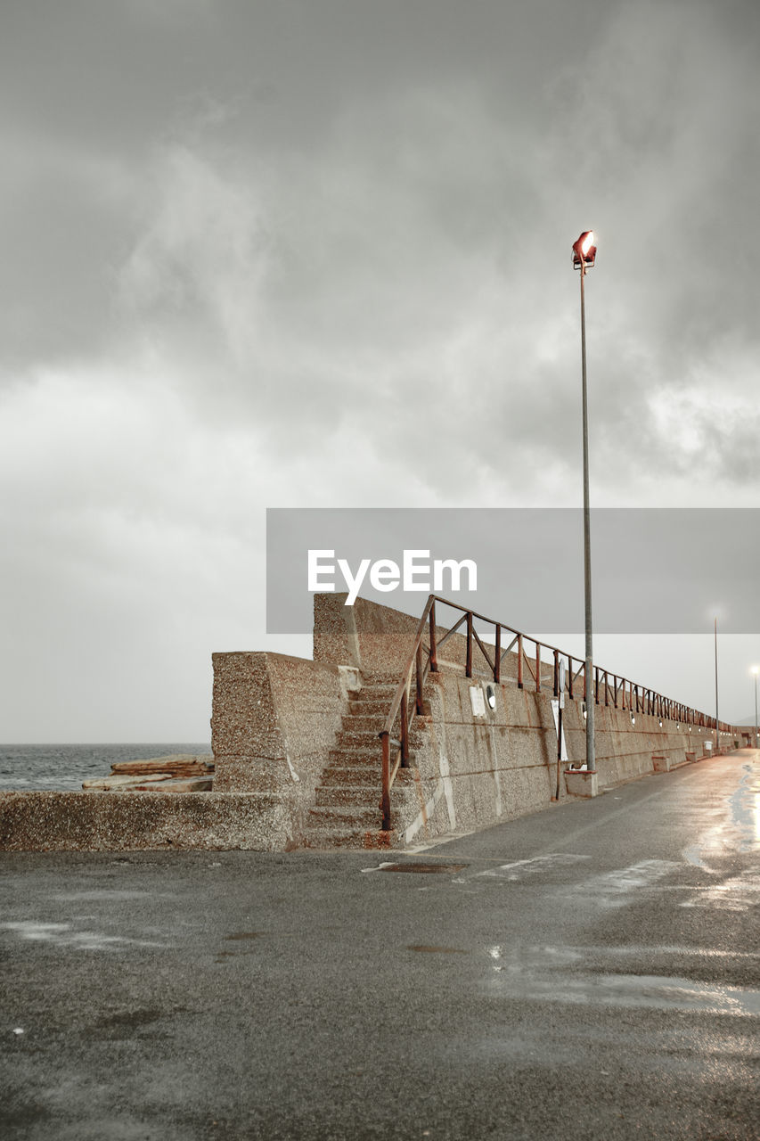 Low angle view of illuminated street light by surrounding wall against cloudy sky at dusk