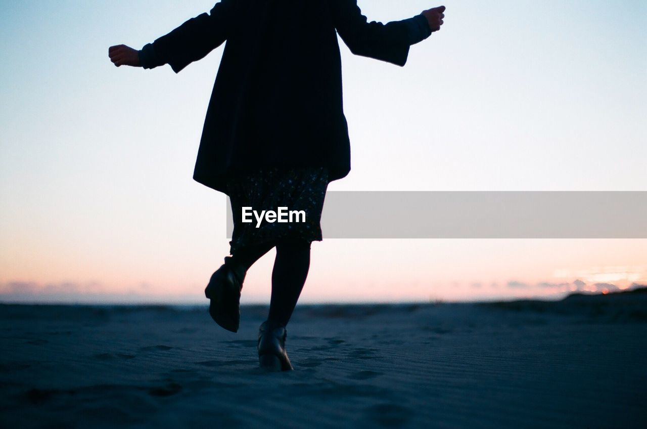 Low section of woman with arms outstretched running at beach during sunset