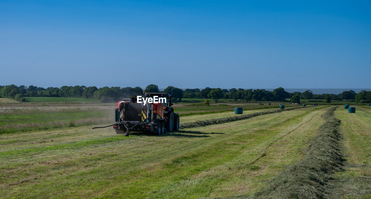 landscape, field, transportation, rural area, rural scene, sky, agriculture, environment, nature, land, plant, farm, grassland, prairie, tractor, plain, grass, mode of transportation, occupation, group of people, adult, agricultural machinery, travel, transport, clear sky, pasture, land vehicle, crop, hill, day, blue, agricultural equipment, scenics - nature, outdoors, farmer, road, men, growth, meadow, vehicle, sunny, machinery, working, dirt road, beauty in nature, harvesting, soil, copy space