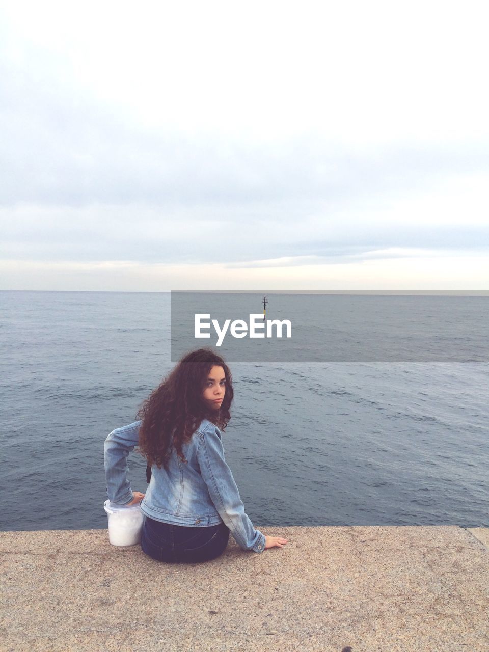 FULL LENGTH PORTRAIT OF YOUNG WOMAN SITTING ON SHORE AGAINST SKY