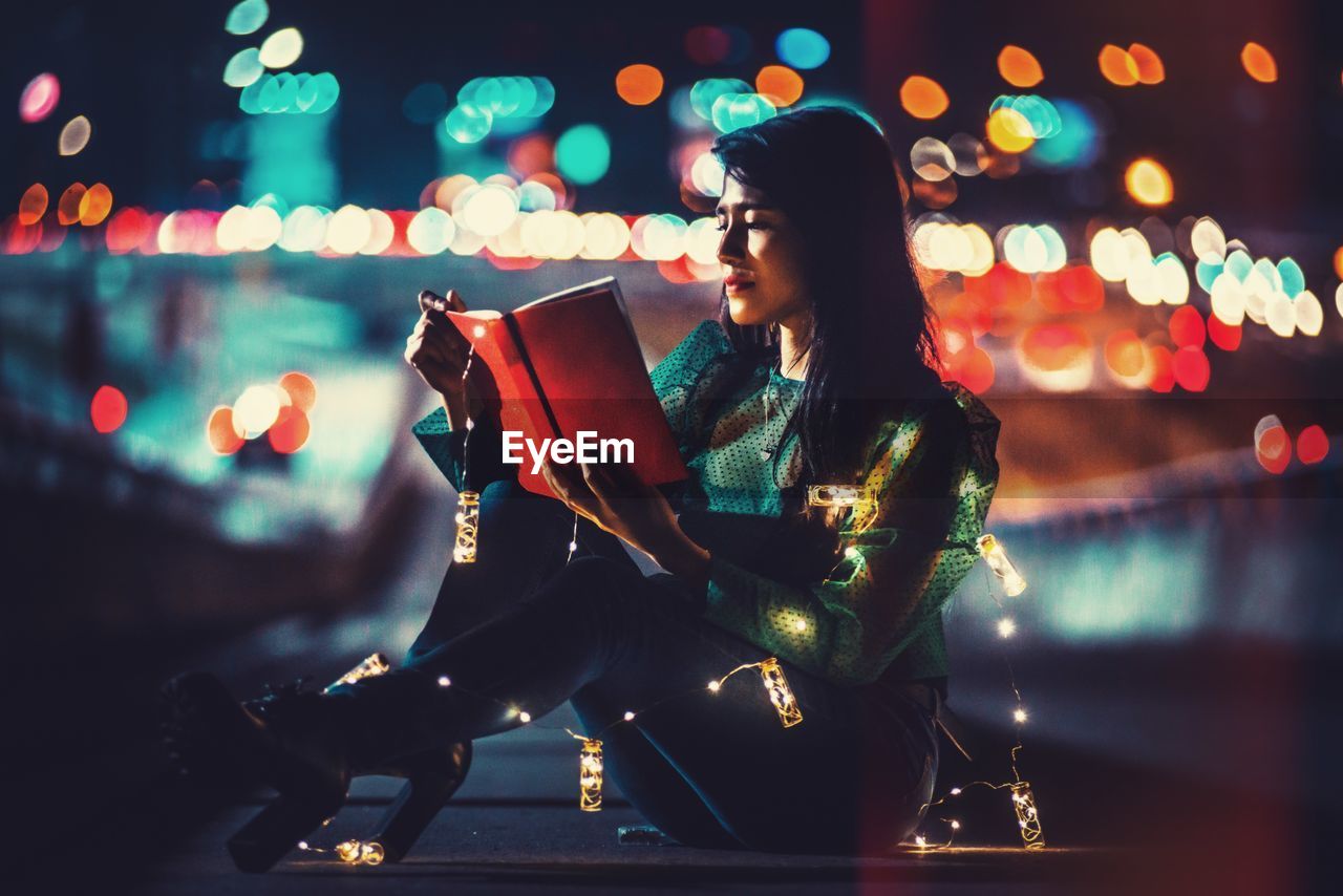 Side view of woman reading books at night near a busy road