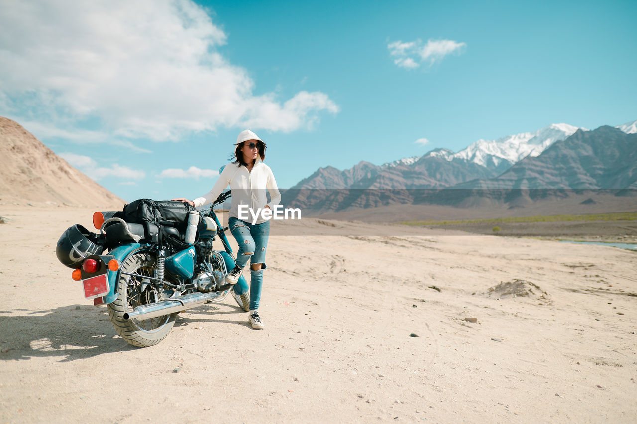 Mid adult woman with motorcycle standing on desert against blue sky during sunny day