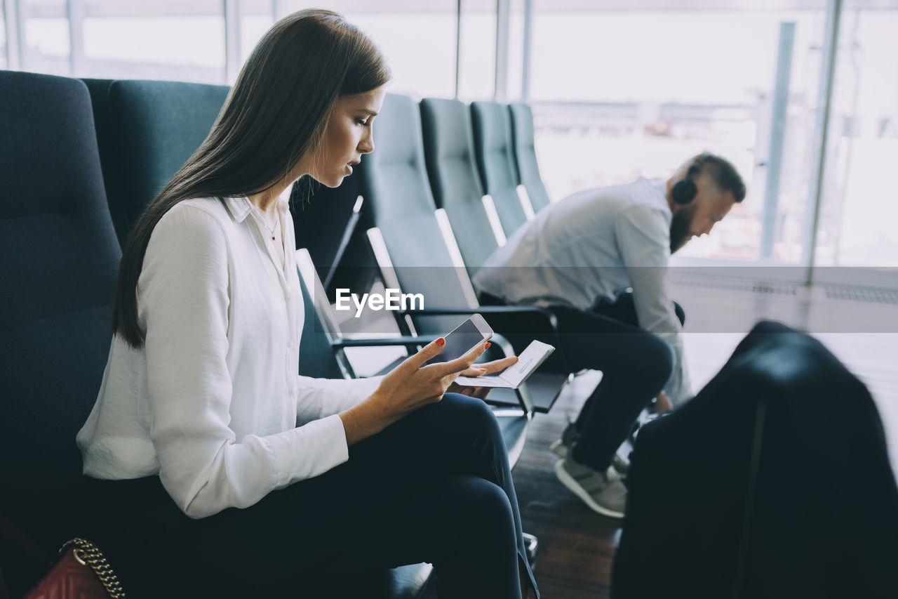 Young businesswoman holding smart phone and passport while sitting at waiting area in airport