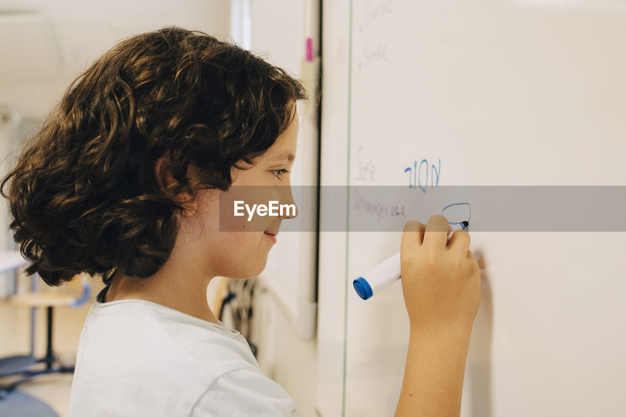 Side view of boy with curly hair writing on whiteboard at classroom
