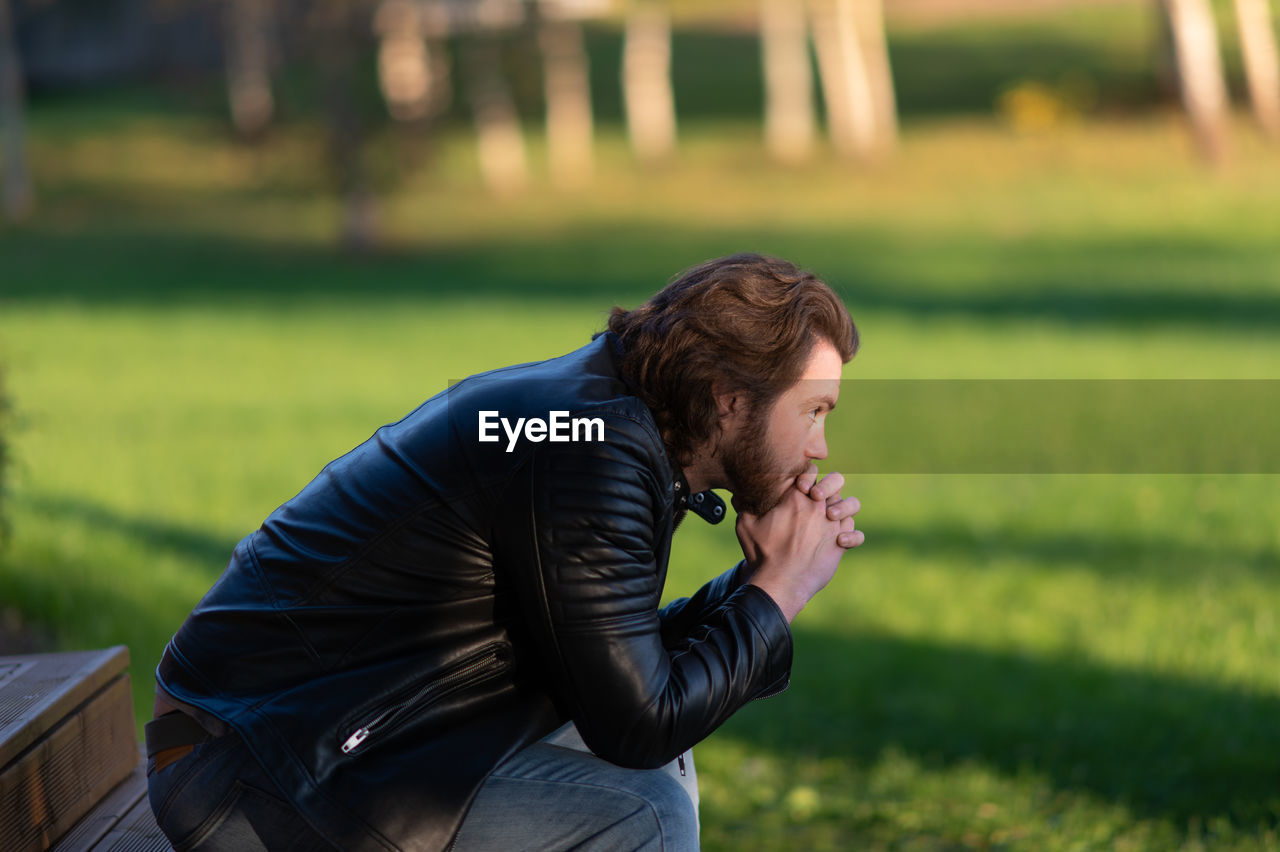 Pensive serious young bearded man wear black leather jacket sitting and thinking outdoors