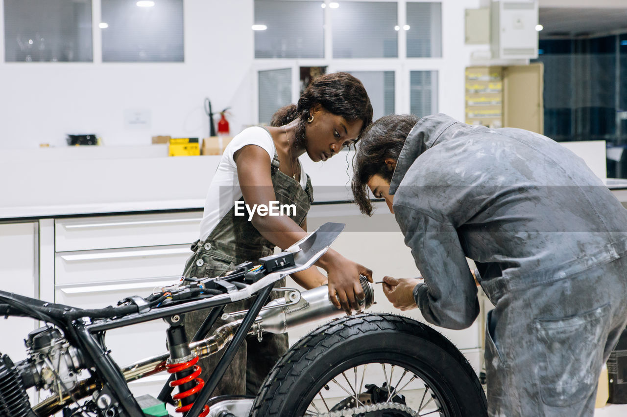 Side view of diverse technicians in workwear repairing muffler of custom motorbike while working together in bright workshop