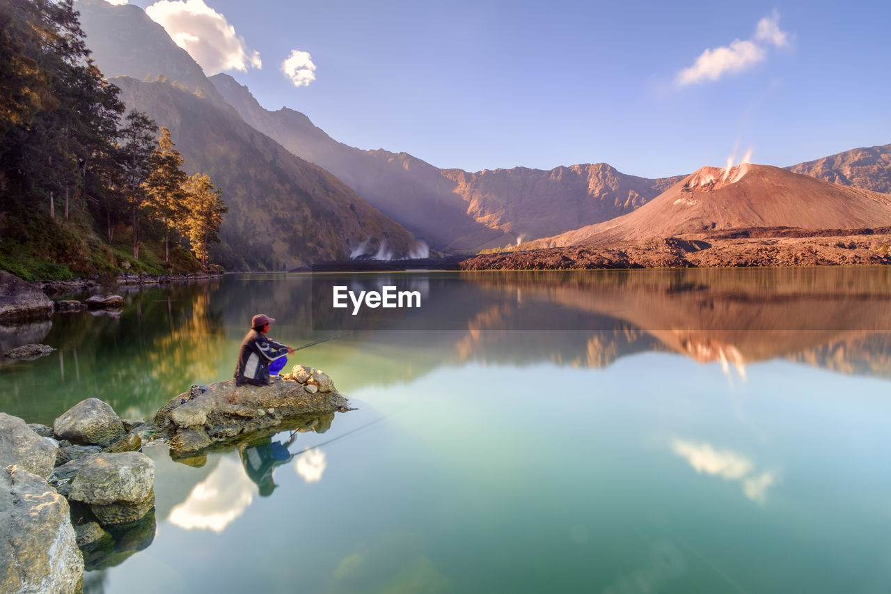 Side view of man fishing in lake while sitting on rock against sky