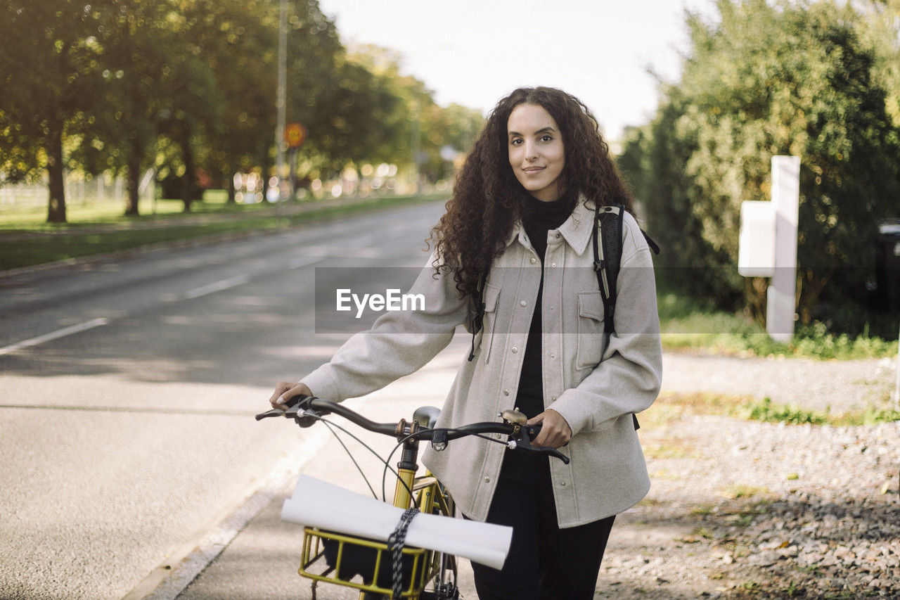 Portrait of smiling female architect wheeling bicycle on sidewalk in city