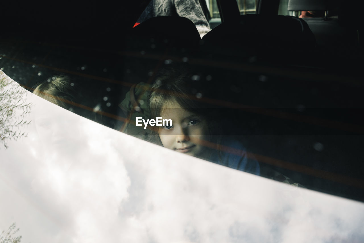 Portrait of girl sitting in electric car trunk seen through rear windshield