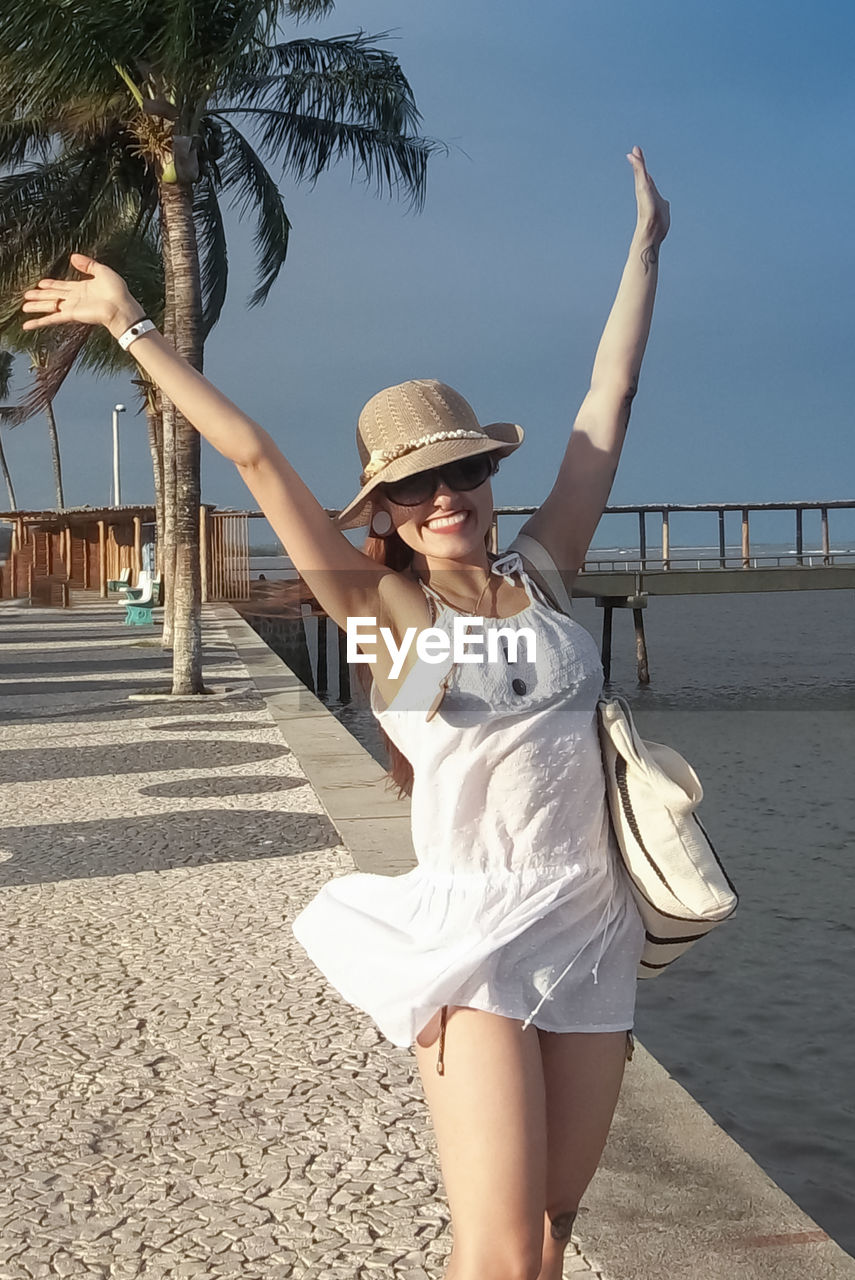 Full length of a young woman, smiling, with arms up in the air over a tropical beach scenery 