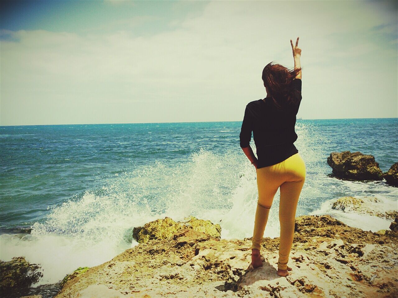 Rear view of woman gesturing peace sign while standing on rock at seaside