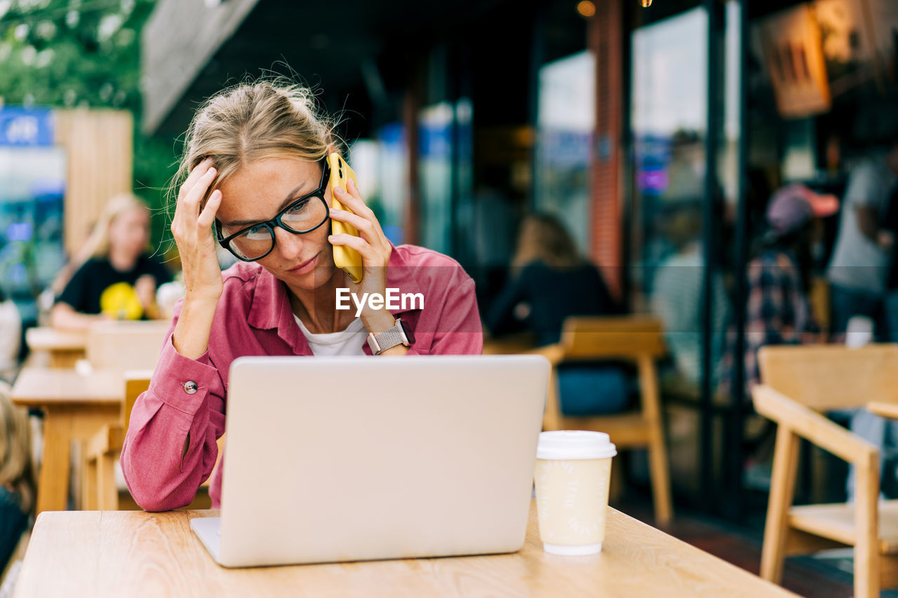 Young working woman talking on the phone and using a laptop while sitting in a coffee shop