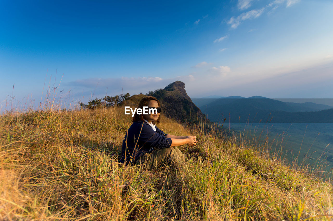 Young woman sitting on grassy mountain against sky