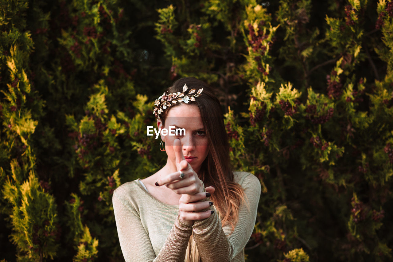 Confident young female wearing trendy hair wreath standing against abundant greenery while pointing finger gun and looking at camera