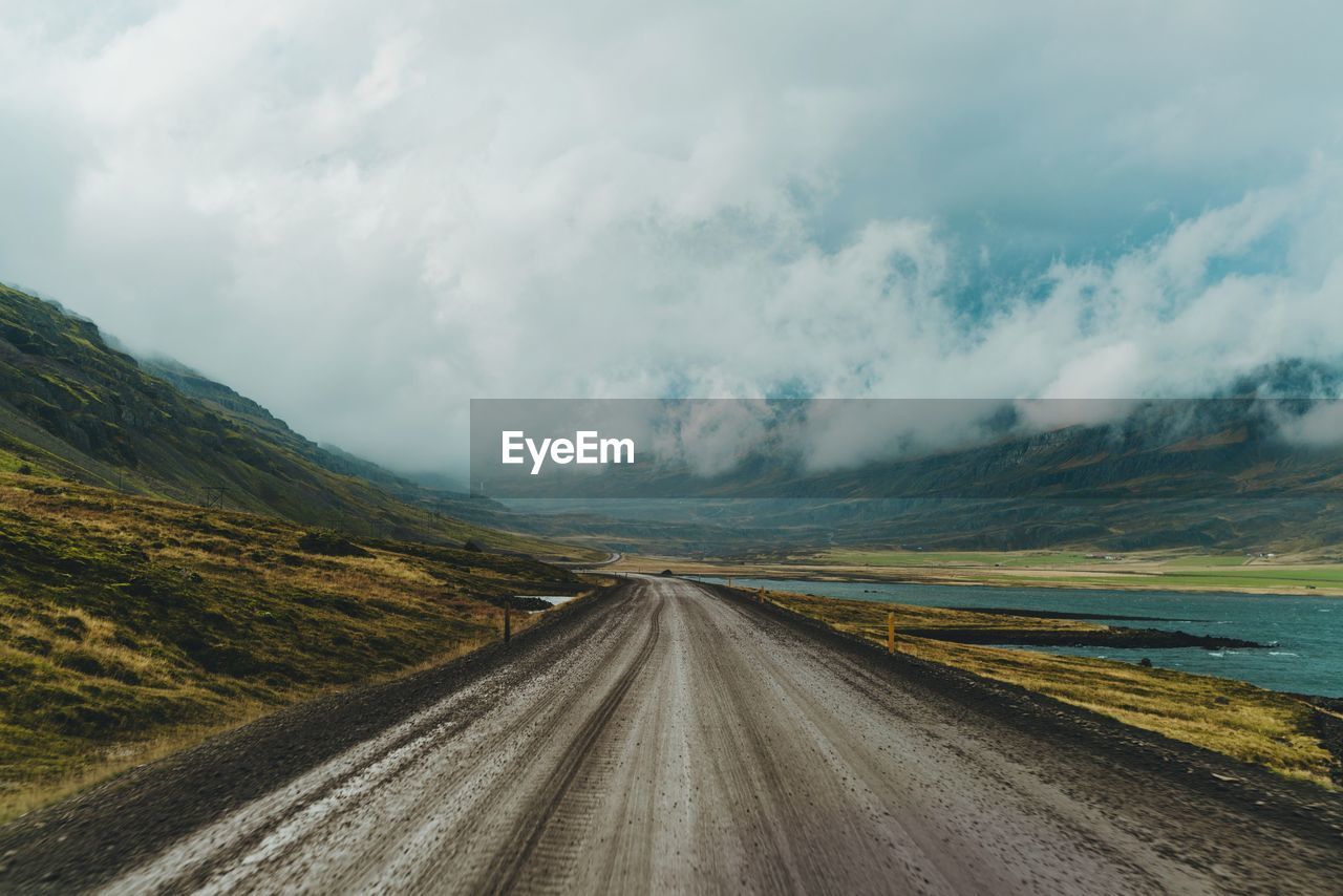 Empty dirt road leading towards mountains against cloudy sky