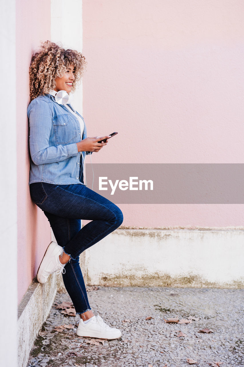 Portrait of smiling hispanic woman with afro hair in city using mobile phone and headset. lifestyle