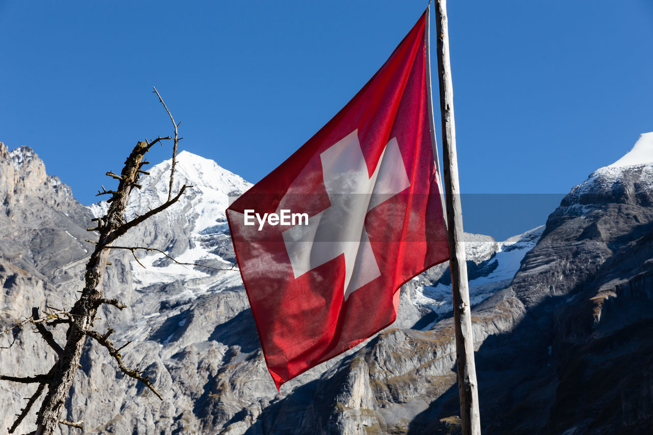 Low angle view of swiss flag by rocky mountains against clear blue sky
