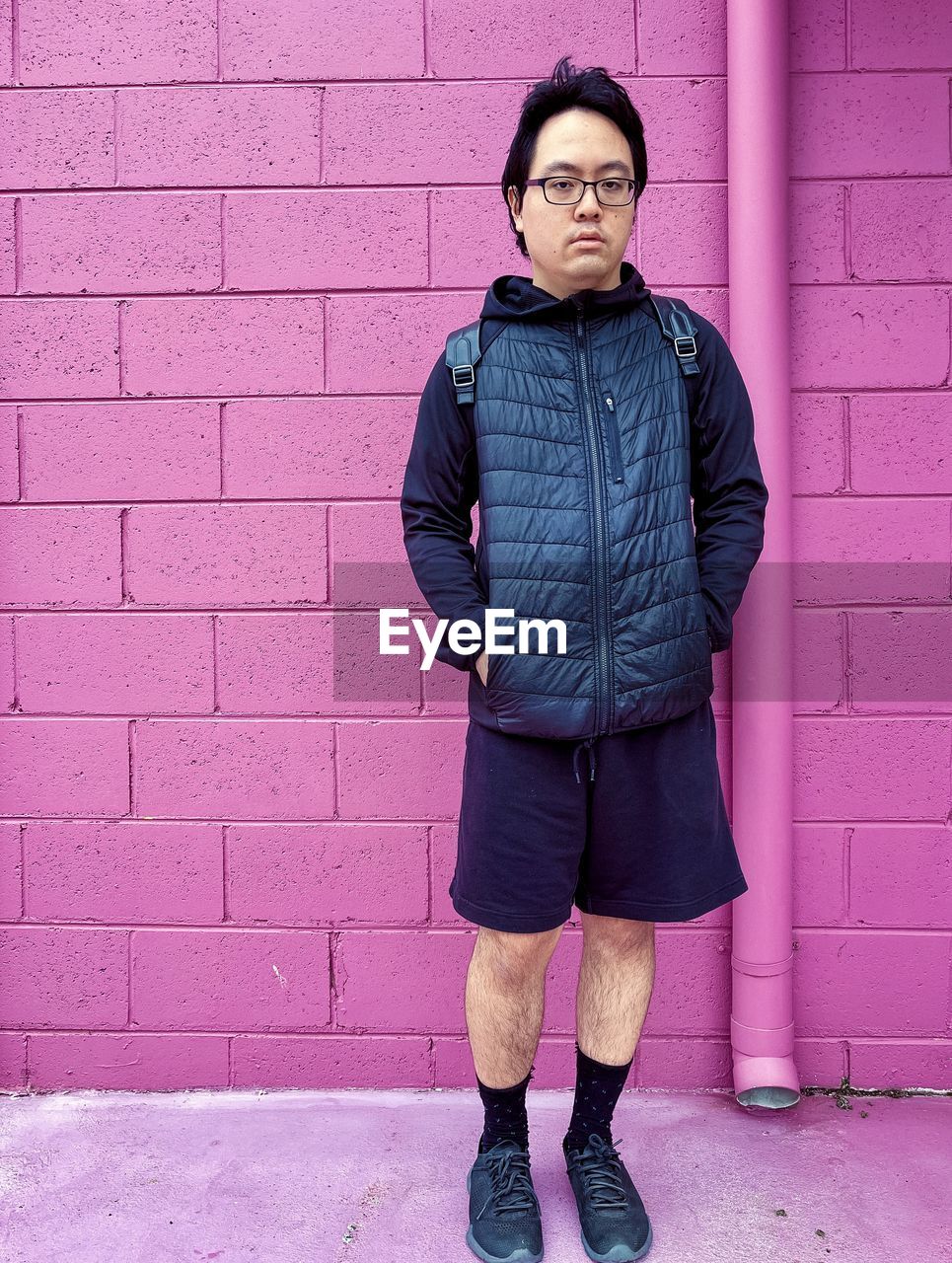 Portrait of young asian man in eyeglasses standing in front of pink brick wall.