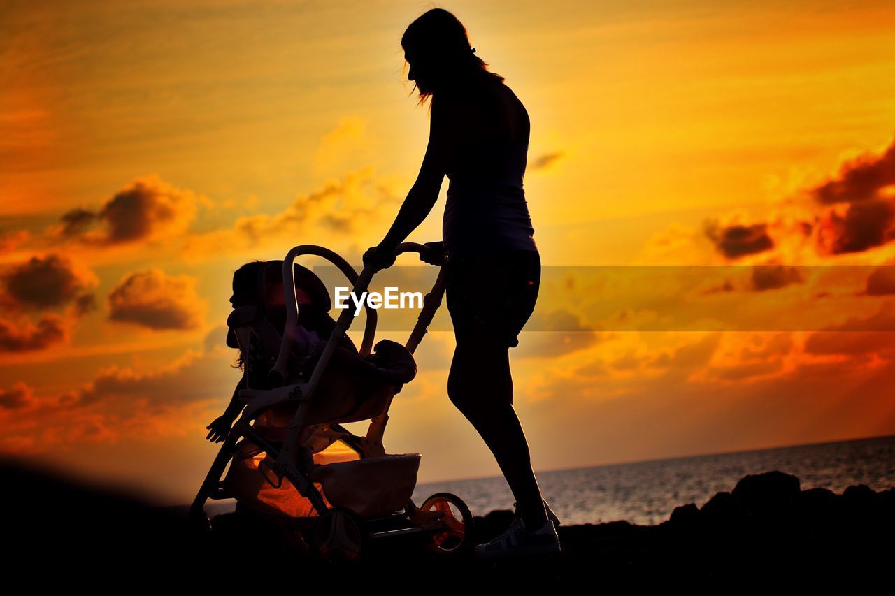 Silhouette woman pushing baby stroller at beach against sky during sunset