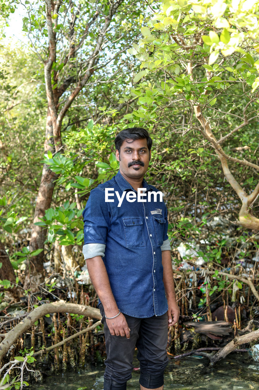 one person, plant, tree, jungle, standing, forest, men, nature, casual clothing, natural environment, front view, looking at camera, rainforest, leisure activity, portrait, young adult, adult, lifestyles, day, growth, land, water, smiling, outdoors, full length, three quarter length, woodland, green, beauty in nature, emotion, happiness