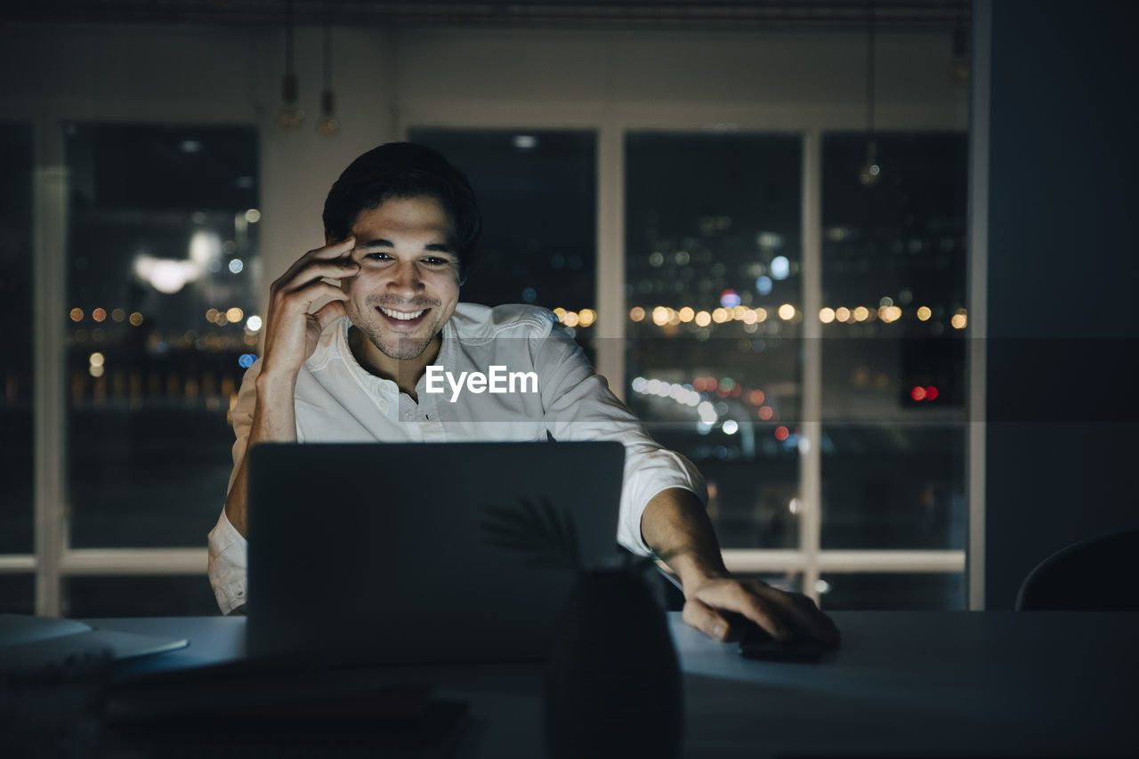 Smiling male professional working late while looking at laptop in dark coworking space