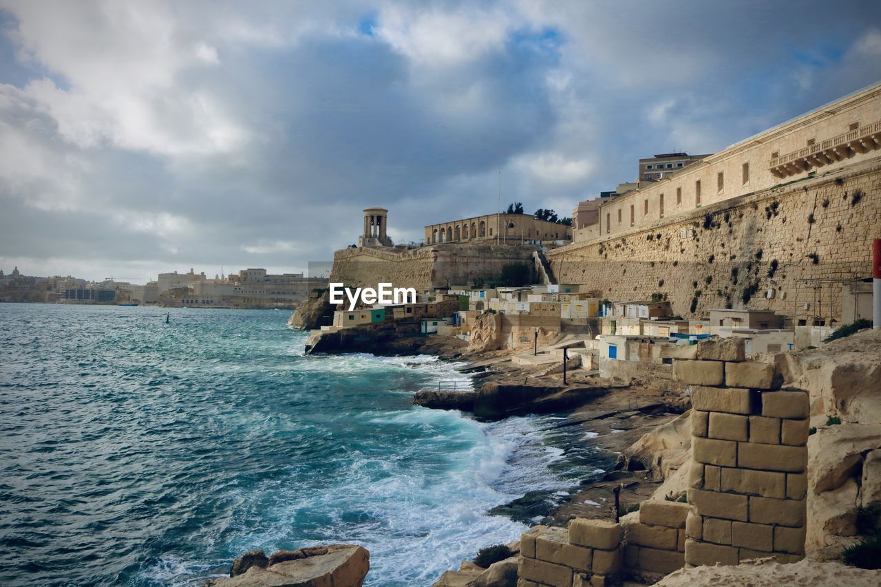 Buildings by sea against cloudy sky