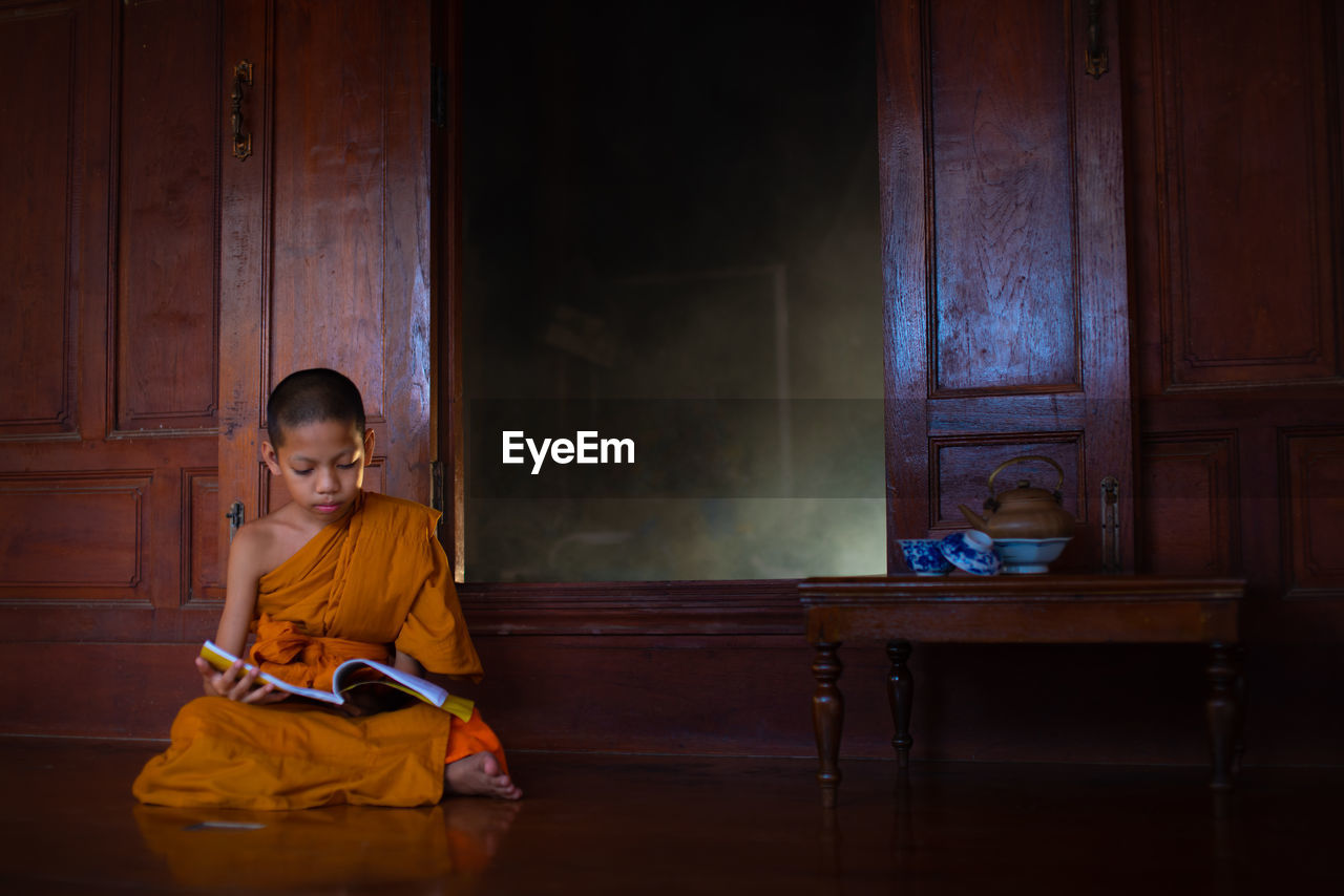 Full length of boy wearing traditional clothing reading book while sitting on floor